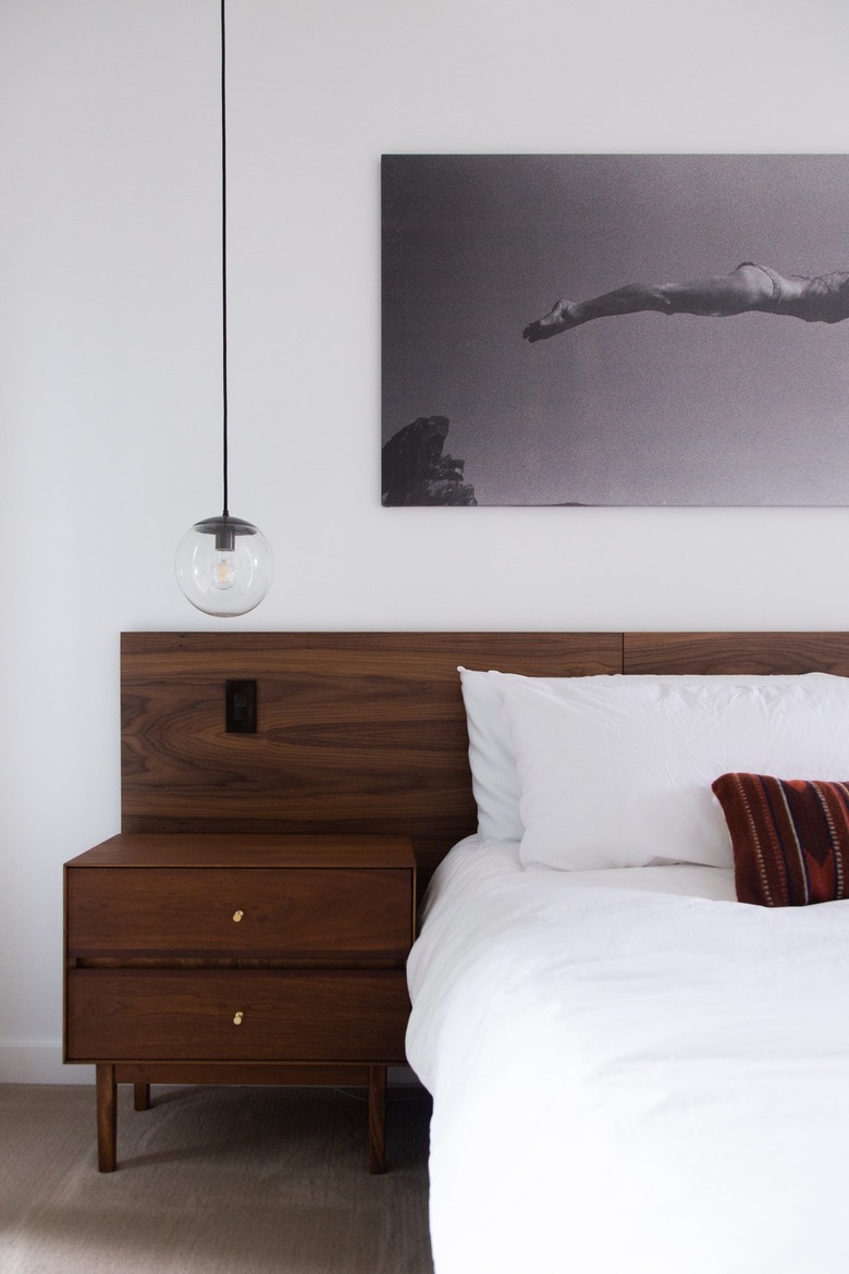 Globe pendant light over a wood nightstand, next to a bed white bedding and a wood headboard.