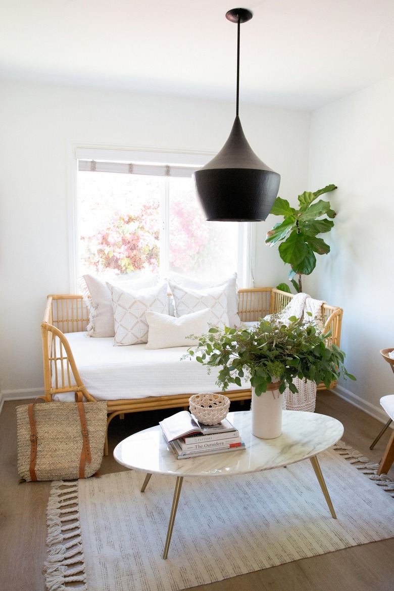 Black pendant light over an oval white coffee table with a vase of flowers and books, rattan bench with white pillows