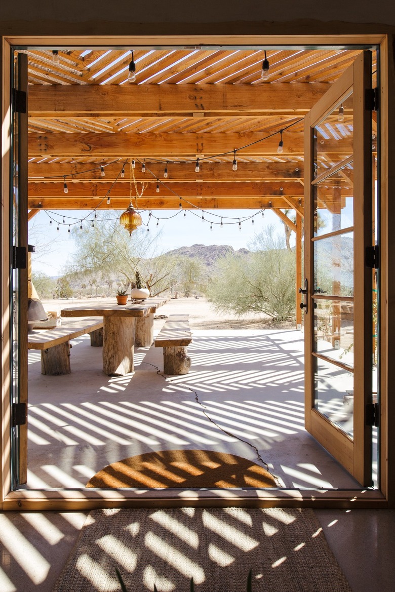 Open French doors, leading to a wood pergola with string lights. A wood picnic table with plants and an ochre pendant light is above.