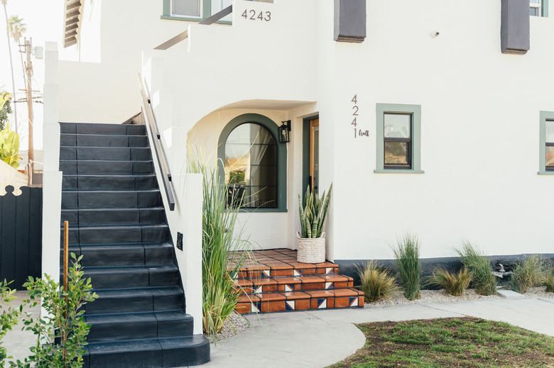 a white Spanish-style multi-family home with an exterior staircase, a curved window, a variety of green plants in a landscape bed with gravel