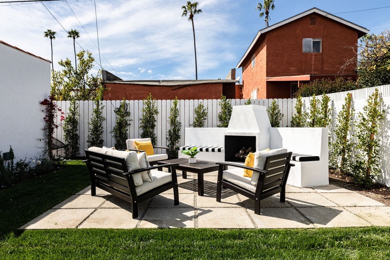 Paver patio with an outdoor fireplace, patio chairs and table surrounded by a white face lined with green trees