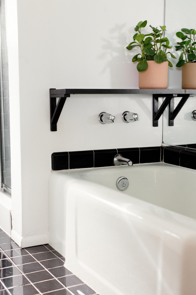 white bathtub with wall-mount silver faucet, black shelf with plant on top, gray square floor tile