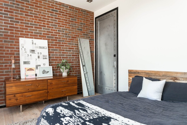 Wood cabinets with lamp, plant, and white diagram board against brick wall next to full-length mirror and black-and-white bed