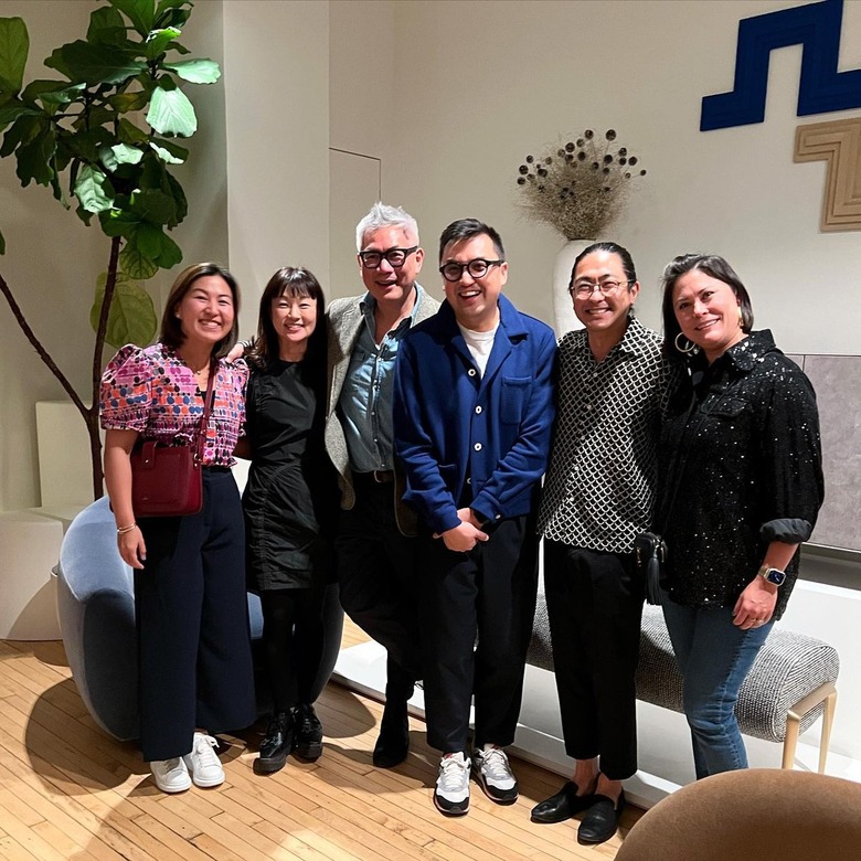 The six founders of the AAPI Design Alliance standing in a row in front of a gray upholstered bench and large plant.