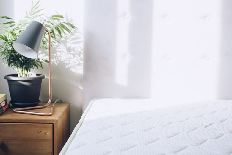 White mattress beside side table with small plant and lamp against white curtains