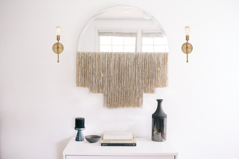 White fringe fringe mirror with gold sconces over dresser with vase