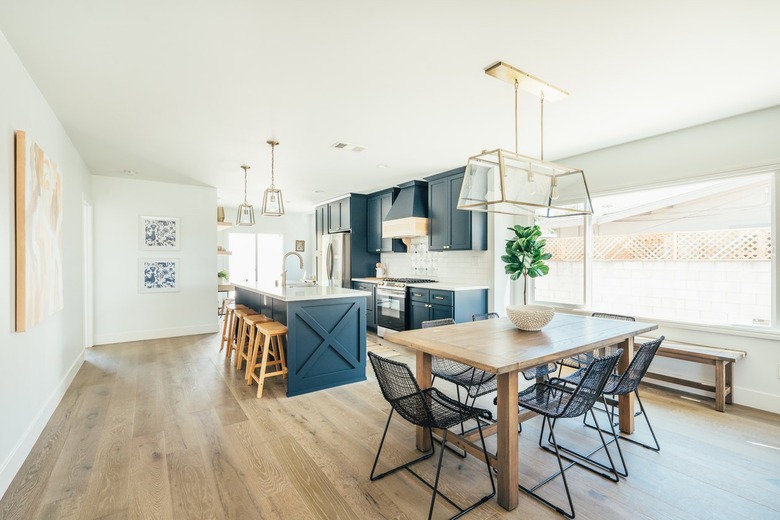 Wood dining room table with black wire chairs and box chandelier. Kitchen with dark blue kitchen island and cabinets.