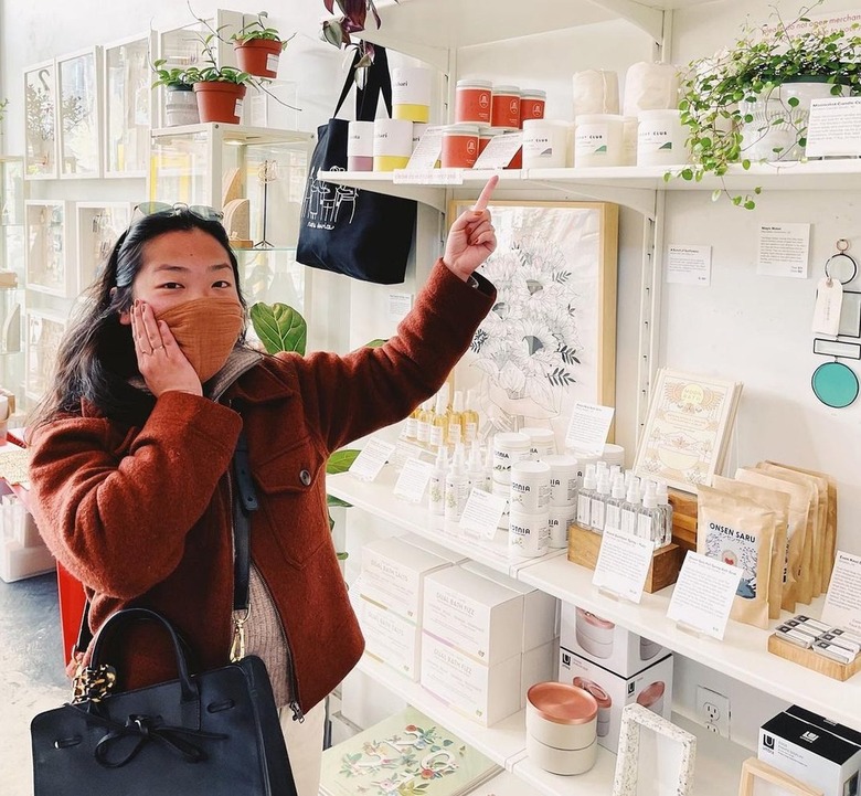 Alice Chen has dark hair and wears a red jacket with an orange face mask while pointing to her candles in a boutique.