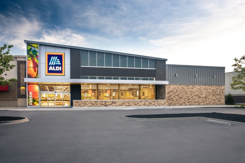 The exterior of an Aldi store. The building has a slanted roof, with the Aldi logo at the top on the right side of the building, with an image of apples on a tree that cover the whole right side of the building, with the word 