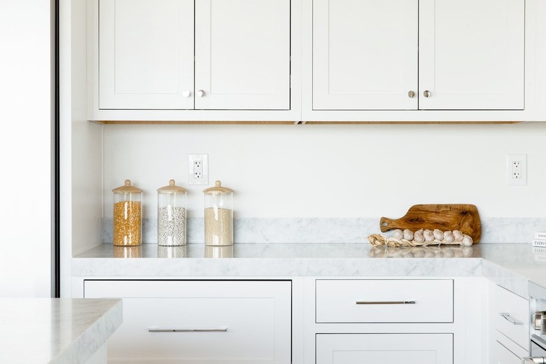 Kitchen counter. The countertop itself is marble, and the cabinets above and below are white. On the counter, three glass food storage containers, a cutting board, and a string of garlic.