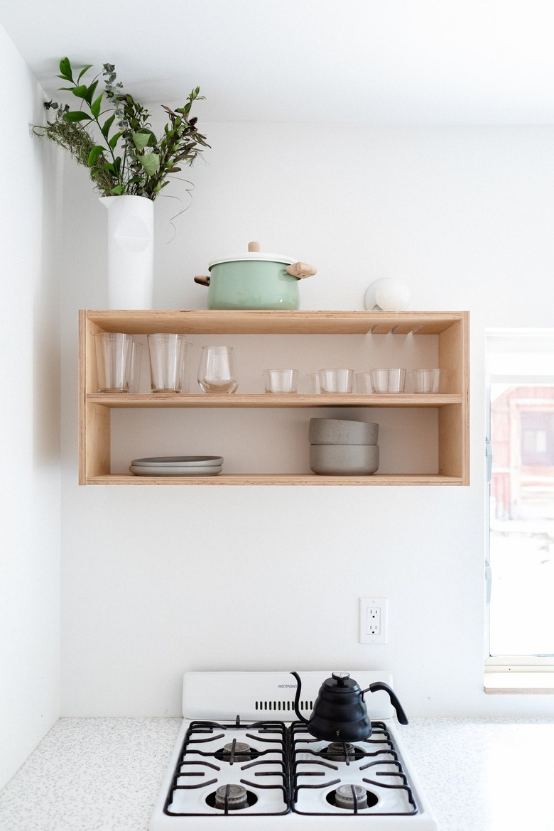 A minimalist white walled kitchen and wood shelving with dishware and plant and apartment size stove