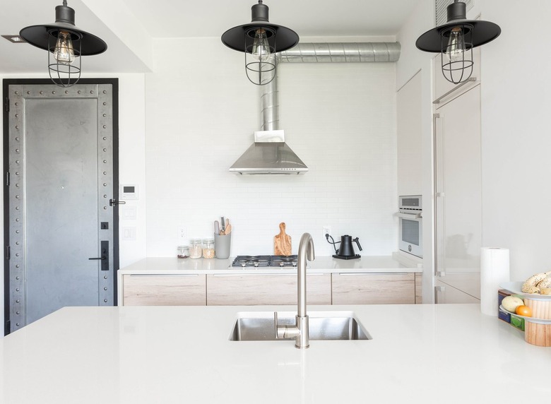 White contemporary kitchen with white countertops, wood cabinets, black stove burners, and silver sink and faucet with black hanging lights