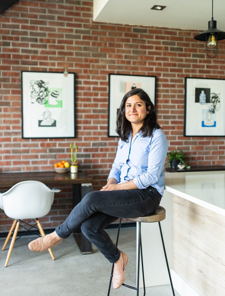 Artist sitting on wood-topped stool in brick-walled gallery space with white wood kitchen island and framed prints