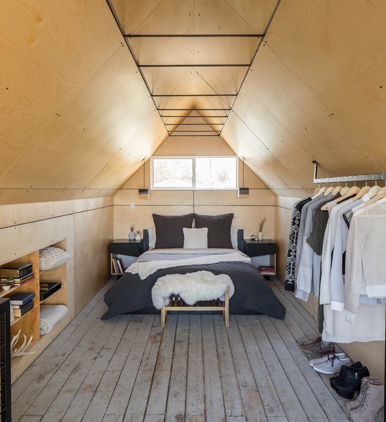 attic apartment with rustic wood floors, plywood walls, wardrobe rack, open shelves, gray bedding.