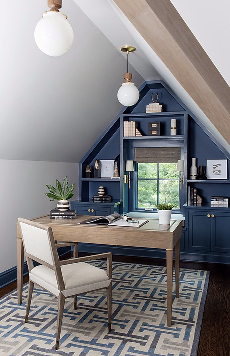 Attic apartment with deep blue wall, built-in shelves, desk, chair, patterned rug, white globe pendant lamps.