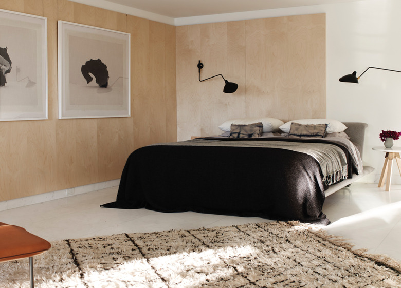 Low platform bed with black and grey linens, black modern sconces black and white large scale photo prints, natural area rug, leather side chair, wooden stool side table, flowers in vase white floors and light wood paneling on walls. Basement Bedroom Ideas