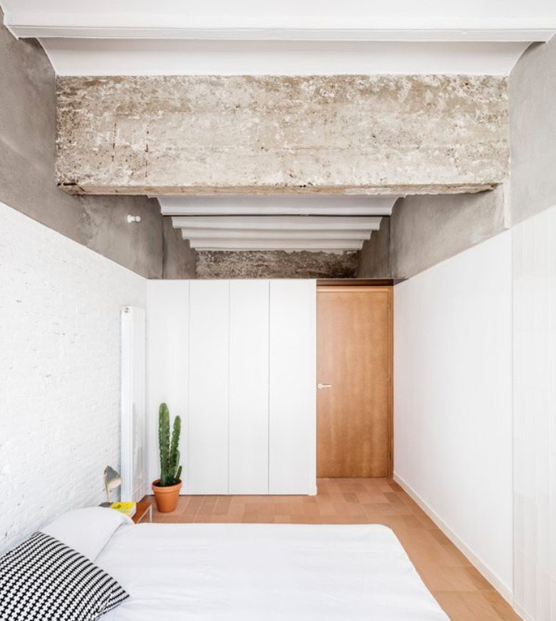 Concrete ceiling, white wood half walls, light wood door and floor, white bed, cactus in terra cotta pot. Basement Bedroom Ideas