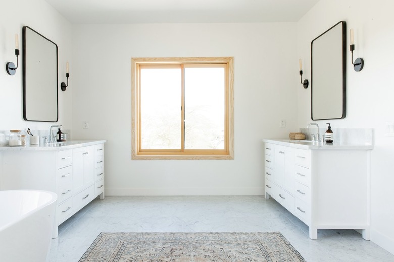 A bathroom with a light wood frame window. Double white vanities, black incandescent light sconces, black frame mirrors are on either side. There is a freestanding tub and a neutral patterned rug.