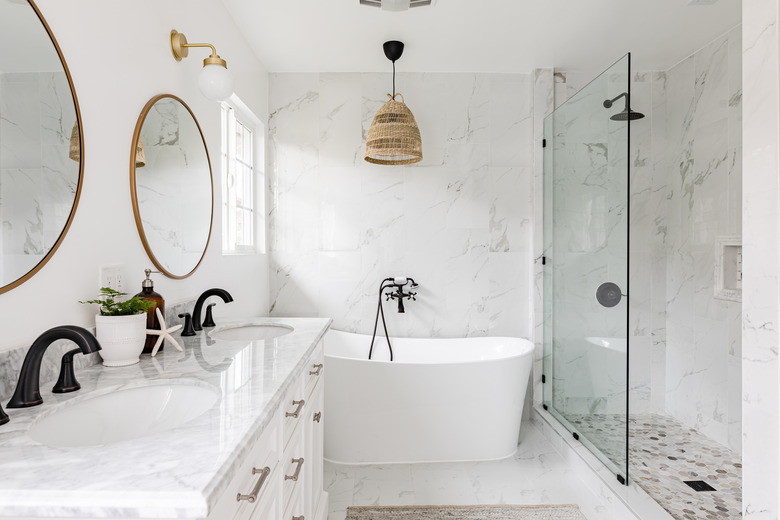 white bathroom, quartz countertop on white vanity, large white soaking tub, glass shower, two round mirrors with gold trim