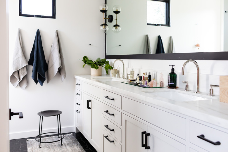 white bathroom vanity with black hardware, double sinks with silver faucet, three hanging towels, black stool, rectangular mirror with black trim