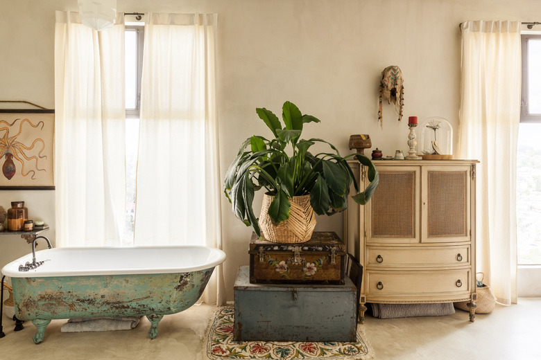 freestanding cast iron tub with vintage distress, trunks stacked on top of each other with a plant on top, a white vintage cabinet is placed between two windows