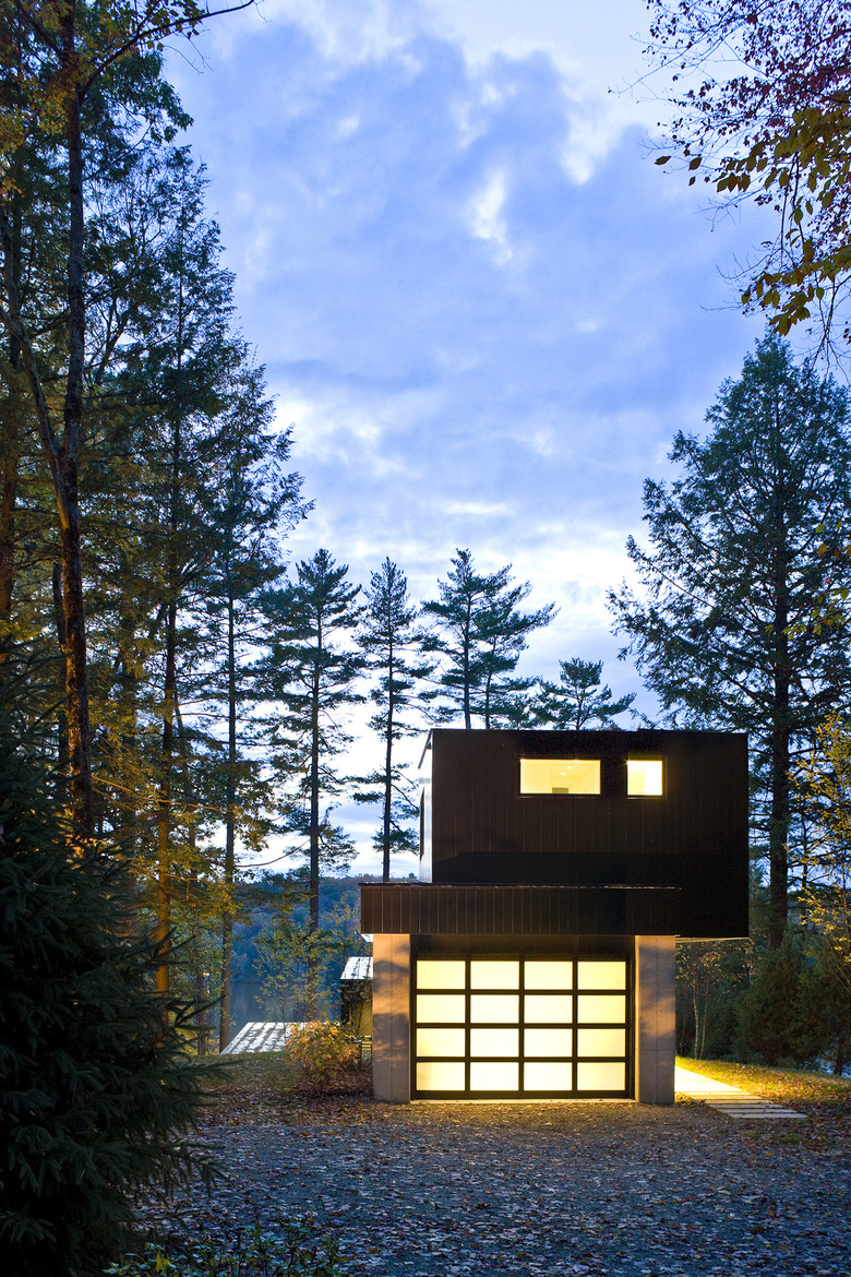 Bauhaus house with second story off-center and lights on in a pine forest