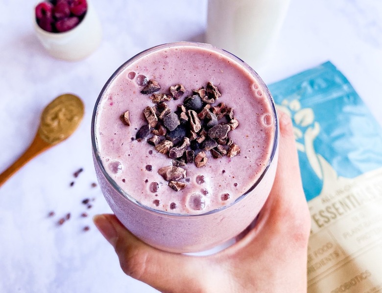 A hand holding a purple smoothie topped with nuts and cacao over a white surface.