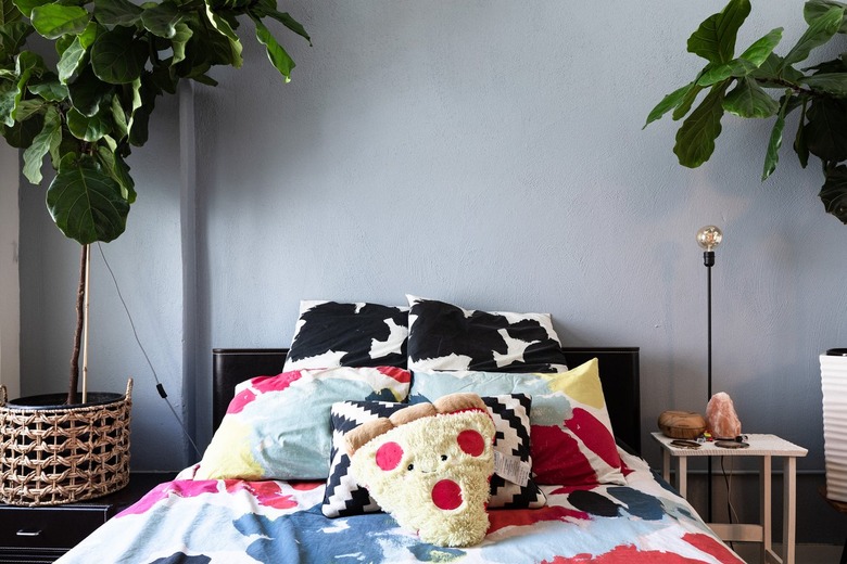 Bedroom with colorful bedsheets, light blue walls, wicker hanging lamp, plants, and side table with small light