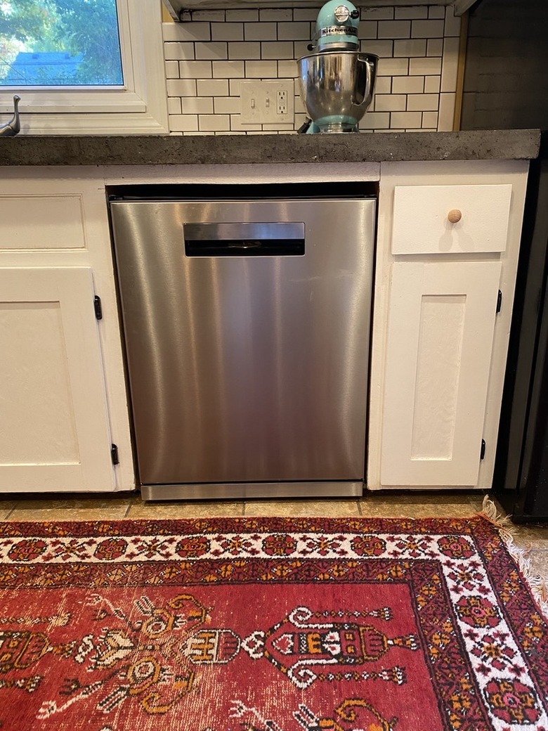 A stainless steel Beko dishwasher built into white cabinets; subway tile backsplash and a red Persian rug