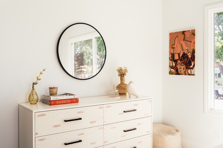 A white dresser with a beige vase and bird sculpture with a round mirrorr and contemporary wall art