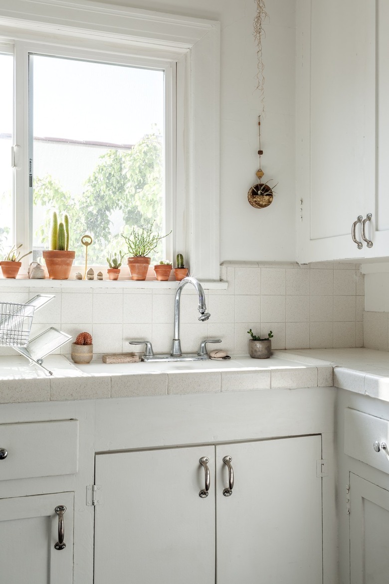 Minimalist kitchen with white cabinets, white tile backsplash, metal faucet sink, and assorted windowsill plants.