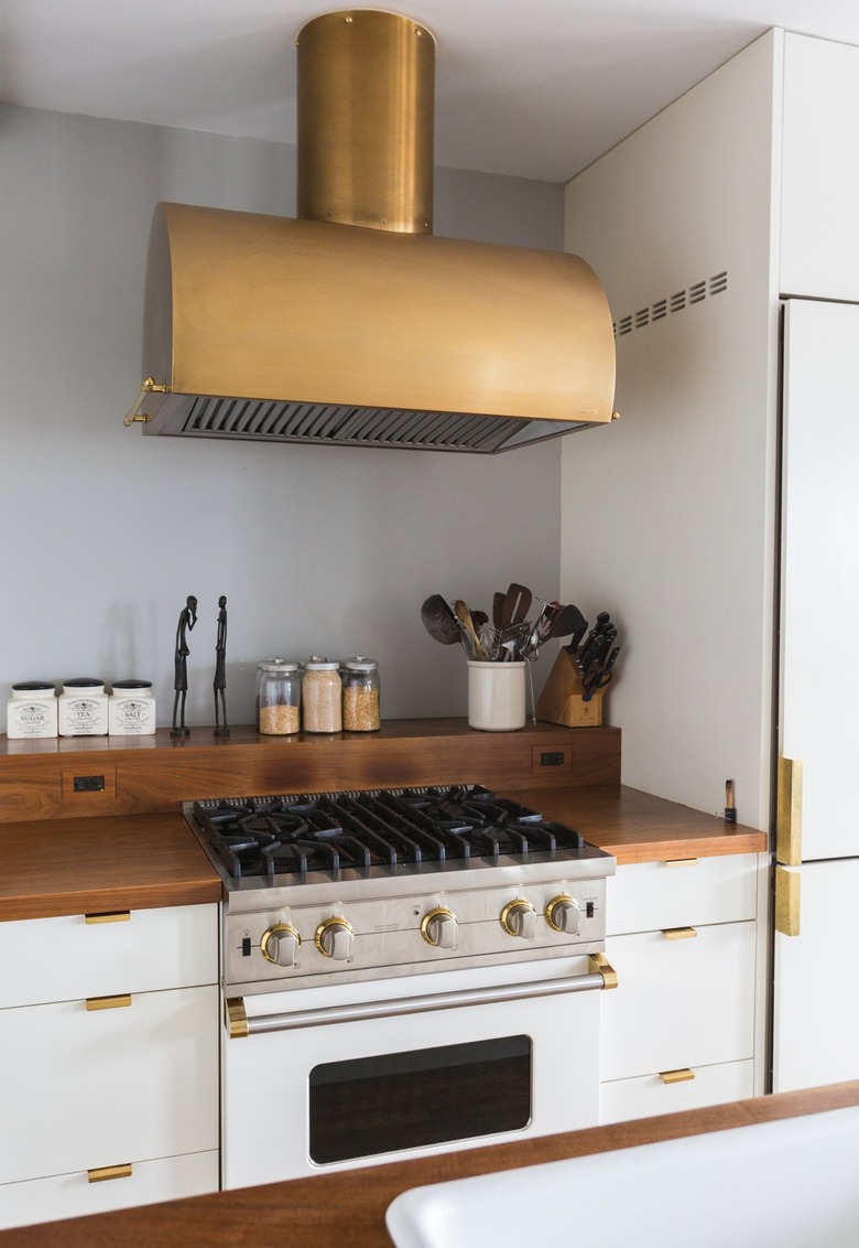 Brass stove hood over a stovetop. White cabinets with a wood counter and brass handles. Containers, sculptures, and utensils on a ledge.