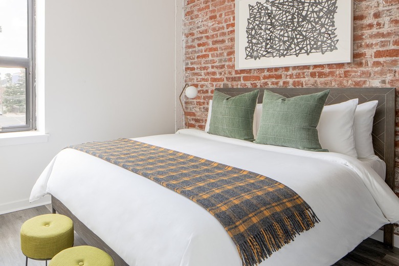 Eclectic bedroom with brick wall, wood headboard, plaid blanket, green pillows, green stools, lamp.