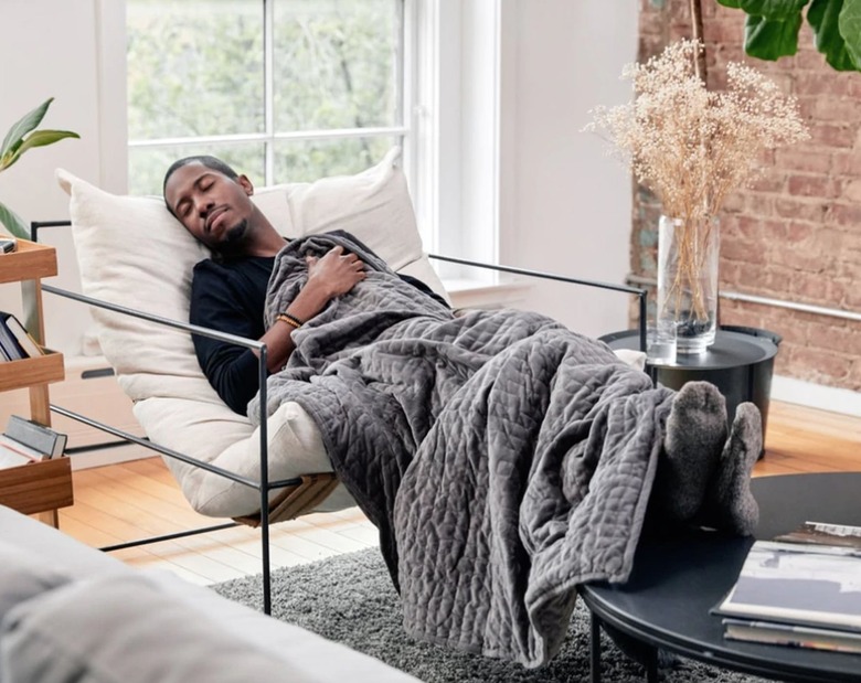 Man napping in chair under weighted blanket, vase, dried flowers, coffee table.