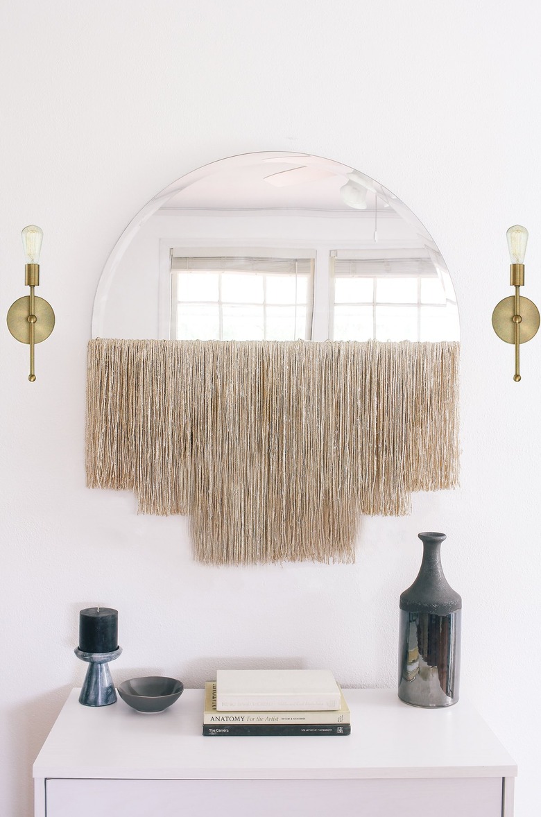 Round mirror with fringe beside gold sconces over white dresser with vase