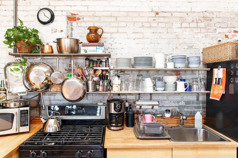 A kitchen with wood counters and white brick walls. Steel shelves with blue-white dish ware, cookware, and a plant.