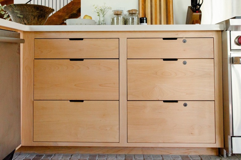 A wood kitchen cabinet with a white countertop. Glass containers, striped wood cutting board, and vases of flowers.