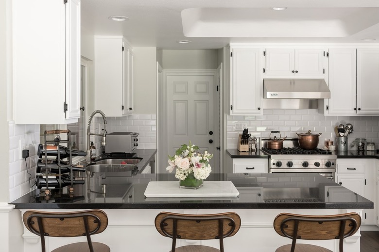 Kitchen with white cabinets, black counters, wood stools, and white subway tile backsplash. A flower arrangement is on a counter.