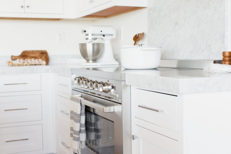 Kitchen counter with white drawers and a white marble countertop. In the middle of the counter, a stainless steel range. A white and grey striped kitchen towel is hanging over the handle over the oven door. A white dutch oven in on the stovetop, and there's a white KitchenAid stand mixer.