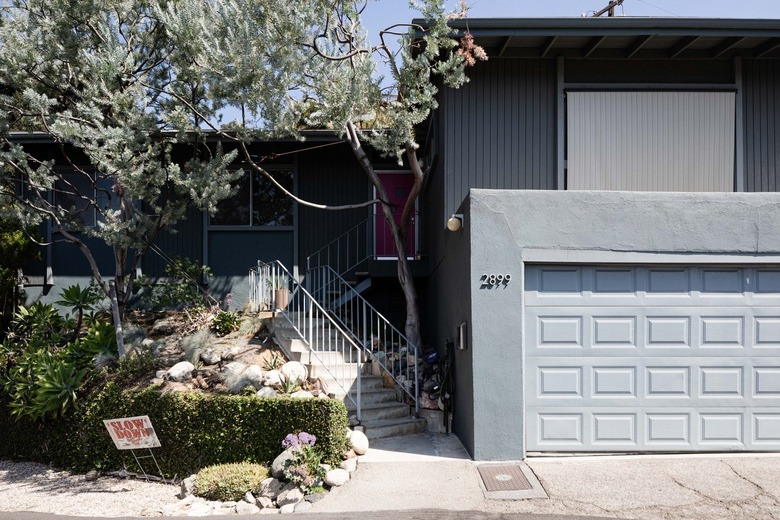 Front of a gray house with a large garage, pink door, front stairs, and trees.