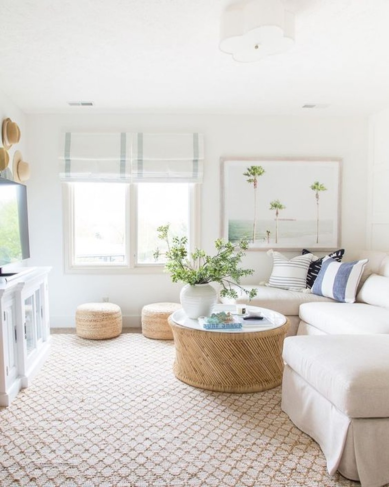 coastal living room with jute rug, off white sectional, wicker poufs, white blinds, white walls, plants, art.