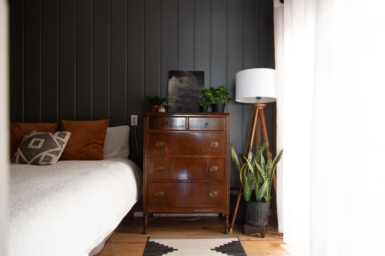 Wood dresser, tripod floor lamp, and black-white southwest rug. Black wall with wainscoting. Bed with brown and gray pillows.