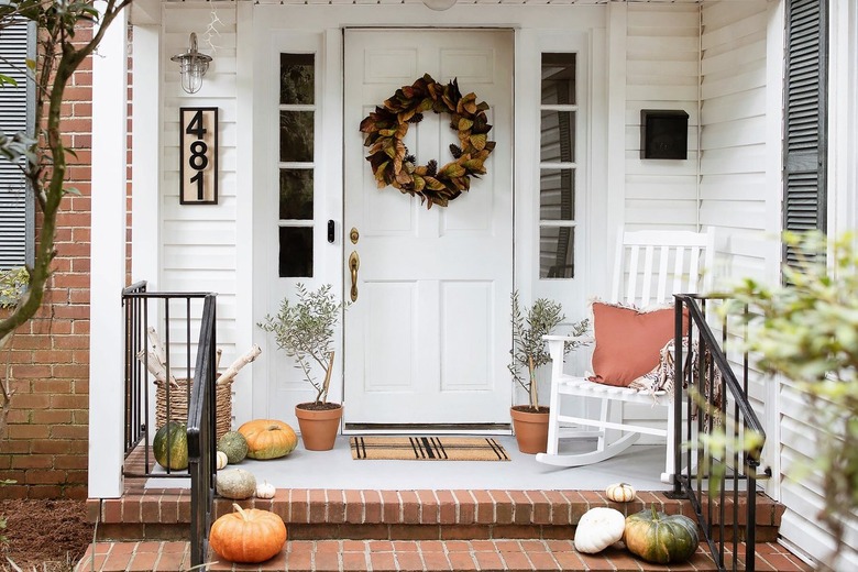 Porch with an autumnal door wreath, tree plants, basket of birch logs, and pumpkins