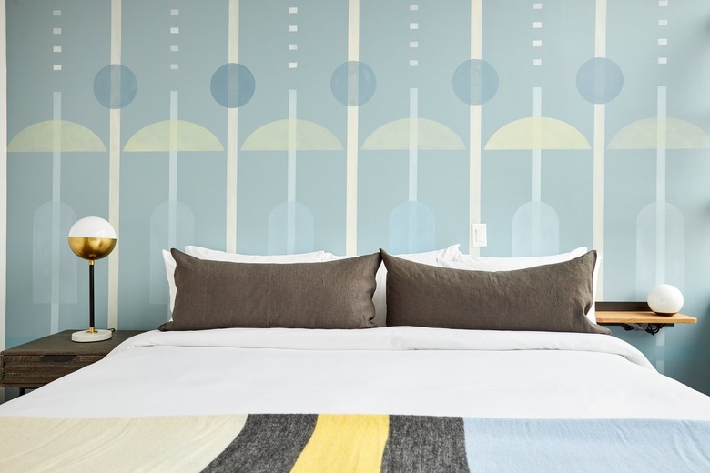 Modern bedroom with geometric wallpaper, brass lamp, white globe lamp, mis matched nightstands, striped blankets.