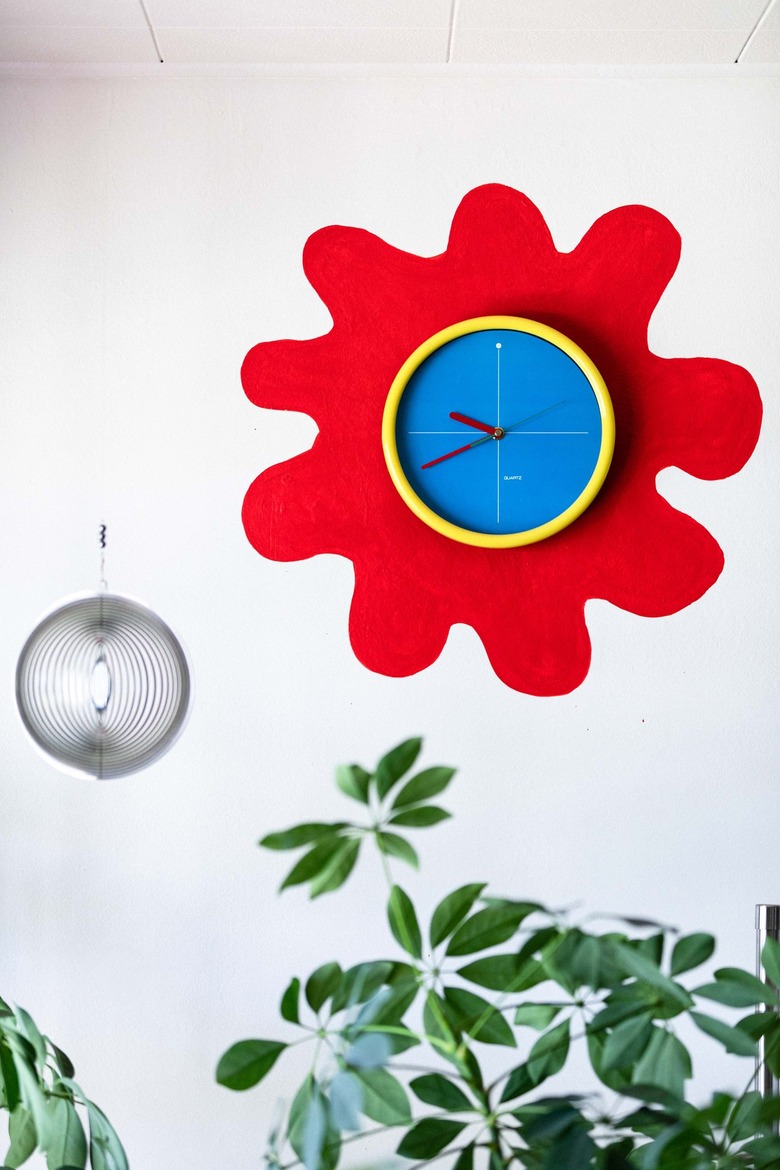 A round blue and yellow clock with a painted red flower on a wall. A globe sculpture is beside. A plant is below.