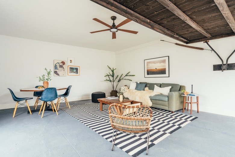 Living room in a California-bohemian style contemporary apartment: cement floor, wood ceiling, ceiling fan, black and white rug, rattan accent chair, plant, coffee table and couch