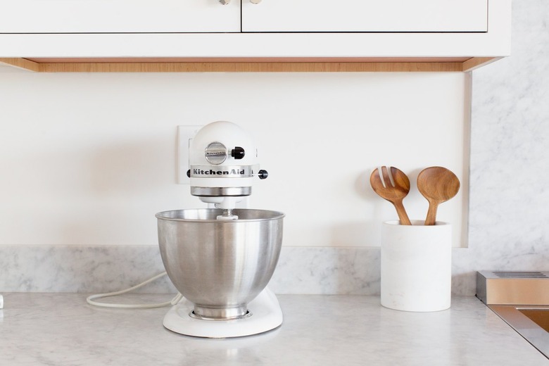 A white KitchenAid stand mixer with a stainless steel mixing bowl on a marble countertop. Next to the mixer is a white utensil holder with wooden salad tongs.