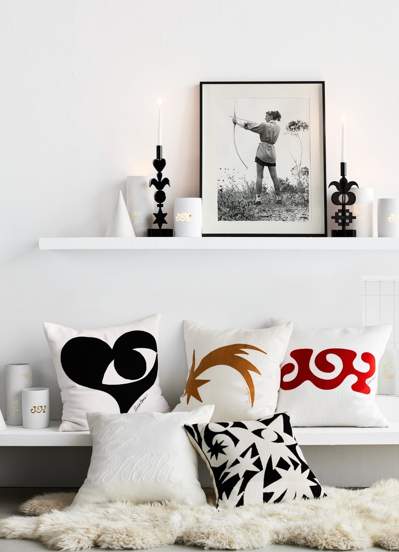 Various Lucia Eames pillows, candlesticks, and glasses displayed on two white shelves.