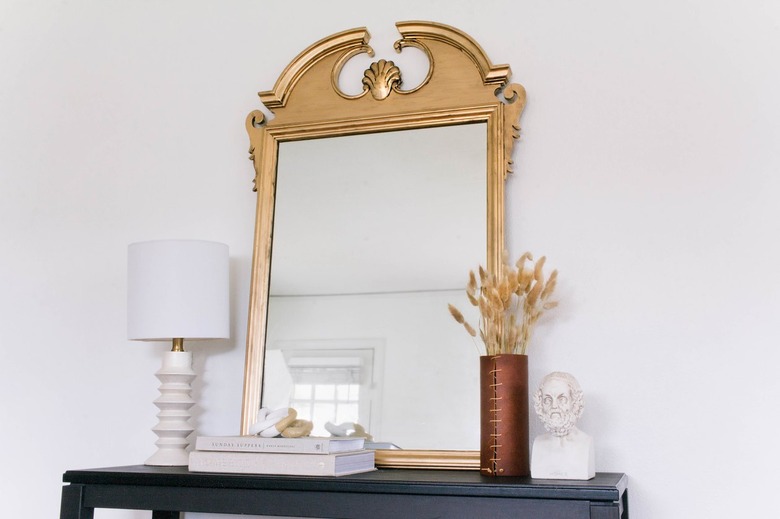 Gold antique style mirror on black side table with lamp, books, vase of dried plants, sculpture bust