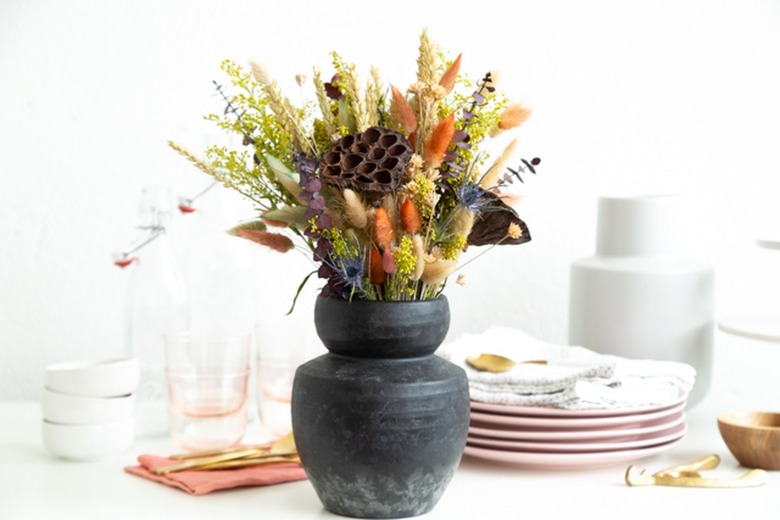 dried flowers in earthy fall colors, including lotus pods and bunny tails dyed burnt orange, in a black earthenware vase surrounded by dinnerware